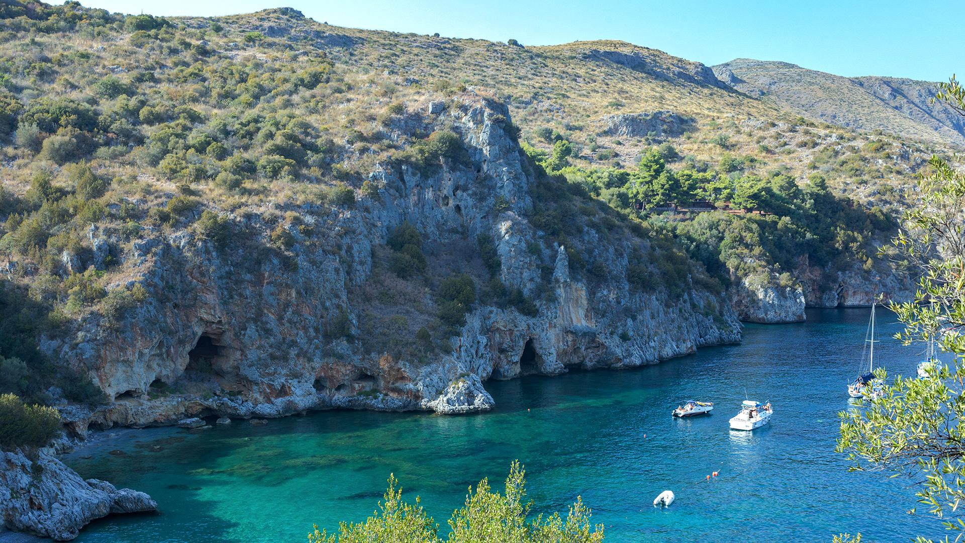 Baia rocciosa con acque turchesi e barche ancorate, circondata da vegetazione mediterranea.