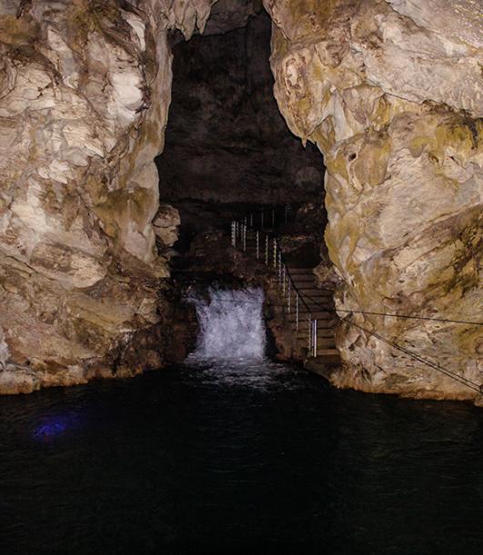 Caverna con cascata interna, acqua scura e scale metalliche lungo le pareti rocciose.