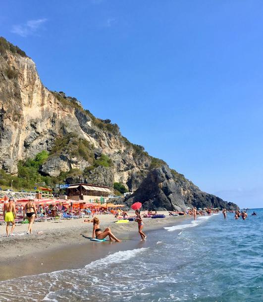 Spiaggia affollata con ombrelloni colorati, mare blu e scogliere rocciose sullo sfondo.