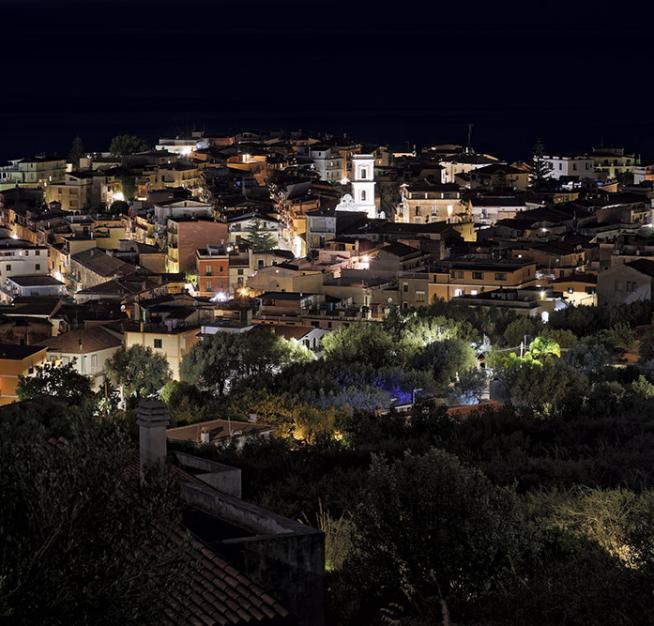 Vista notturna di un pittoresco borgo illuminato, circondato da vegetazione.