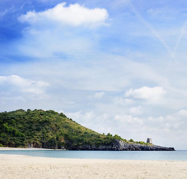 Spiaggia sabbiosa con mare calmo, colline verdi e una torre in lontananza.