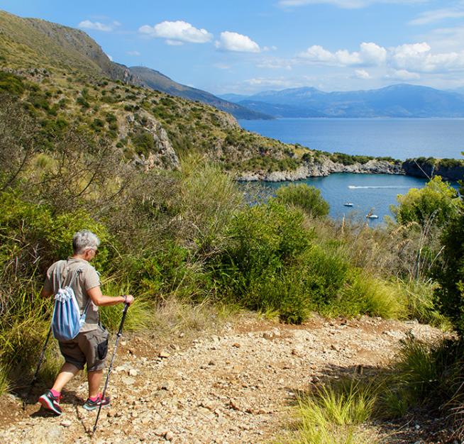 Escursionista su sentiero costiero con vista mare e montagne sullo sfondo.